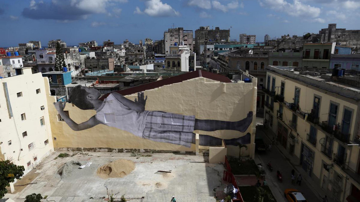 Large mural of a child lying down, painted on a building's wall amidst urban skyline under a partly cloudy sky.