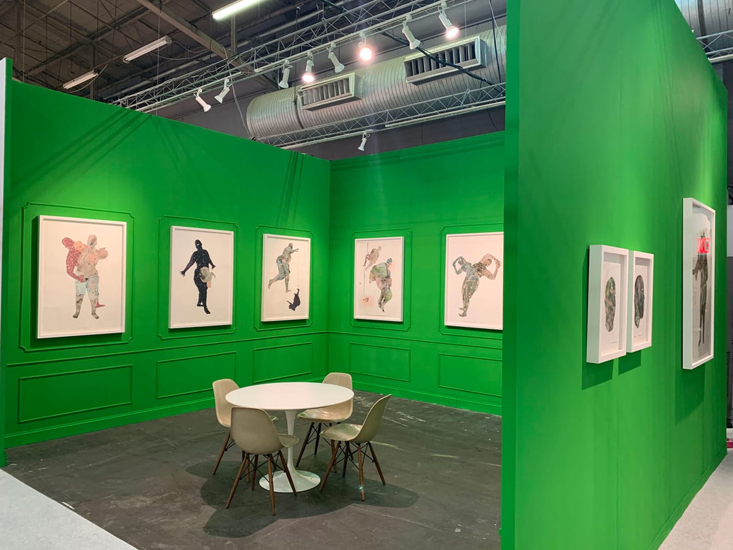Art exhibition room with green walls displaying framed artwork and a round table with chairs in the center.