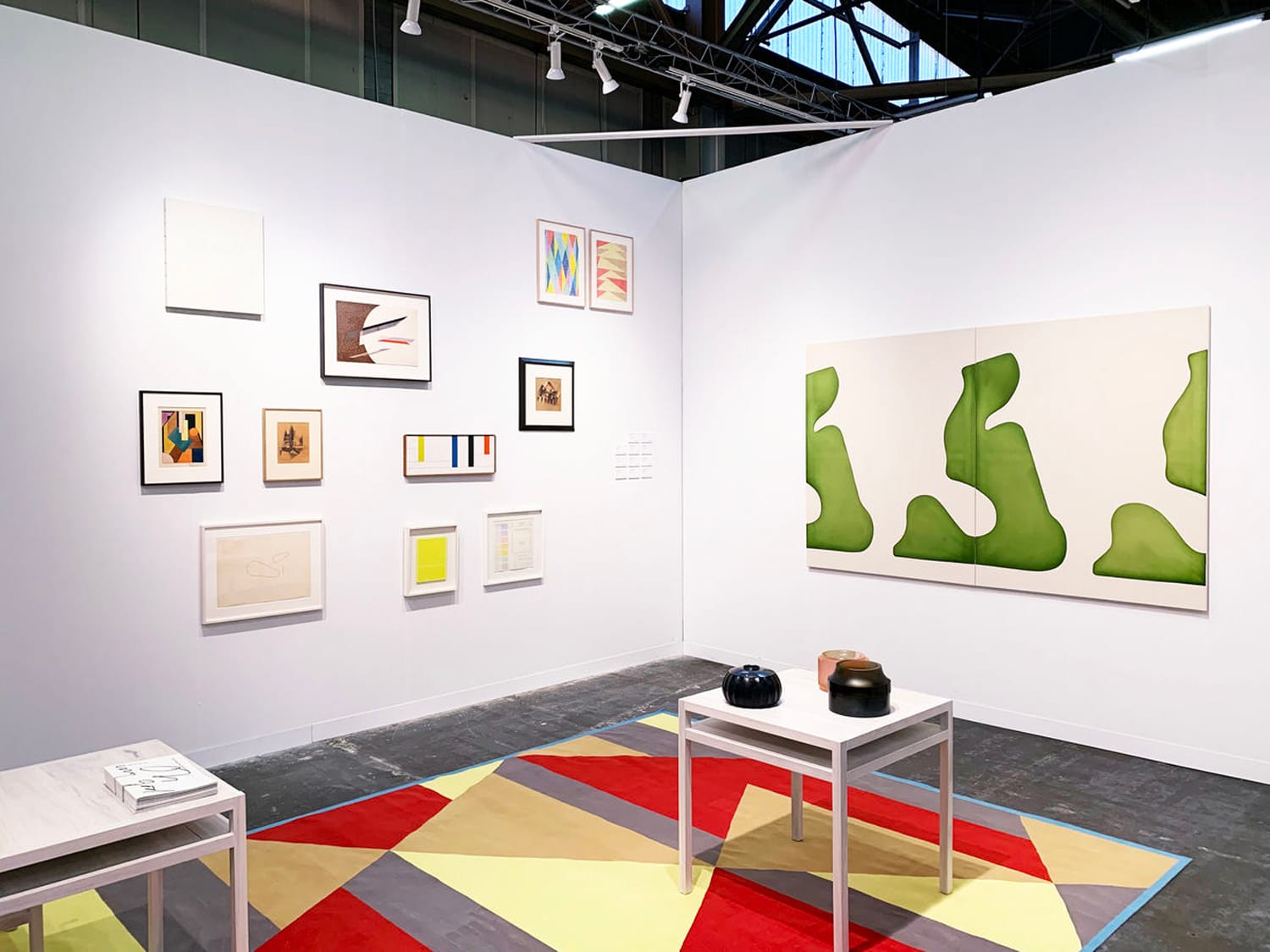 Art gallery display with abstract paintings on white walls, colorful geometric carpet, and white tables with black vases.