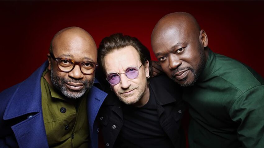Three men posing together against a red background, wearing glasses and different colored shirts.