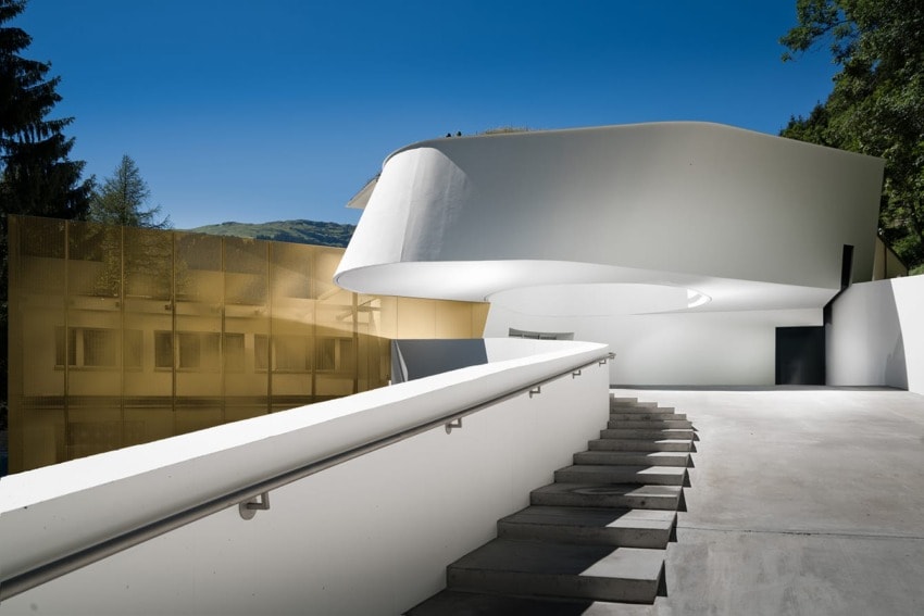 Futuristic white building with curving architecture, glass facade, and steps leading to entrance under clear blue sky.