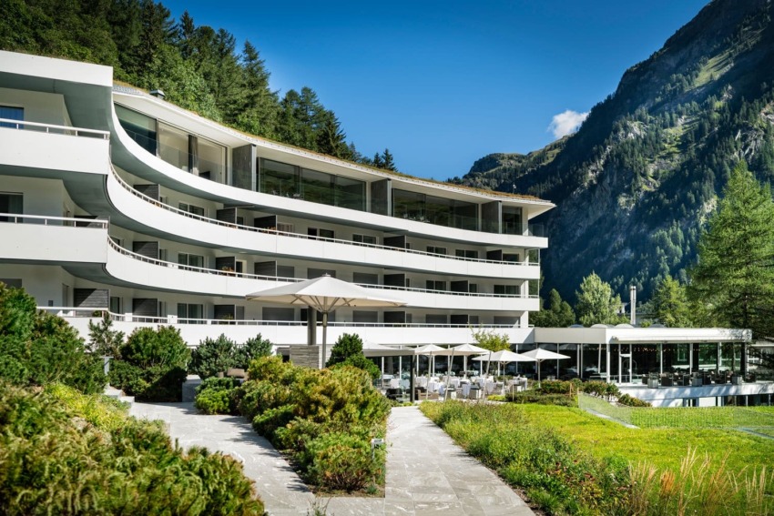 Modern hotel building nestled in a lush mountain landscape with blue sky and outdoor seating area.