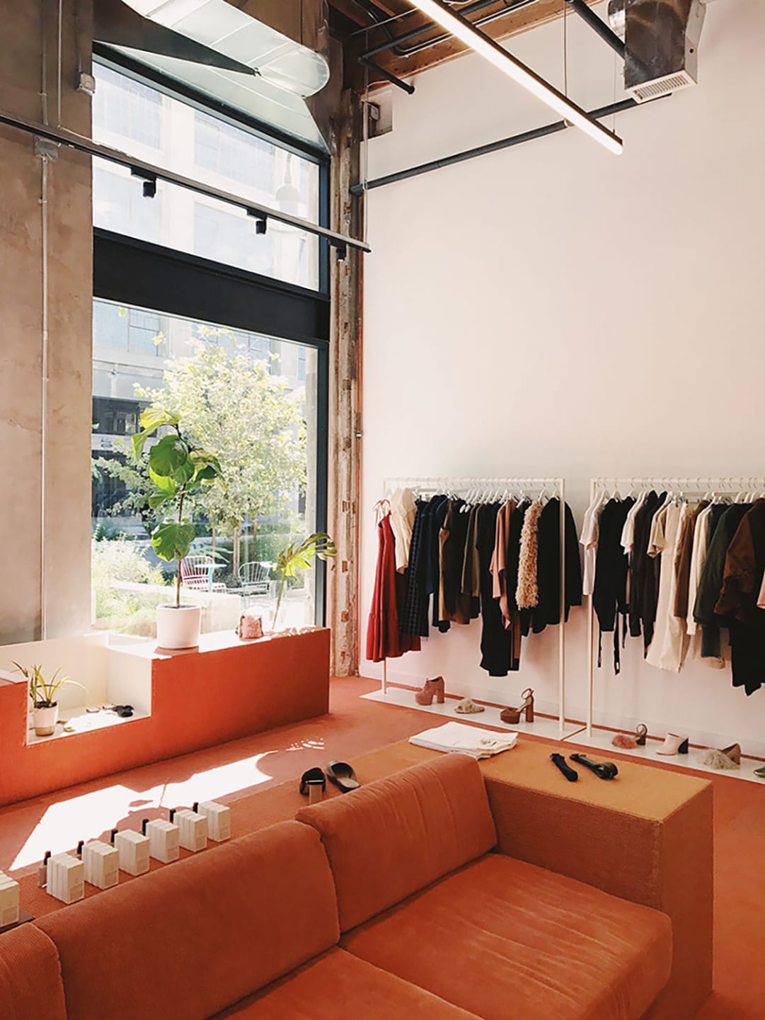 Modern boutique interior with orange seating, clothing rack, large window, greenery, and sunlit atmosphere.