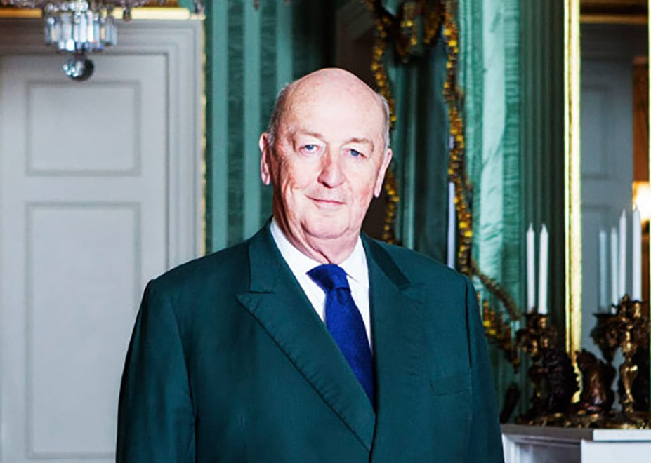 Elderly man in a suit standing in an elegant room with green walls and a chandelier.