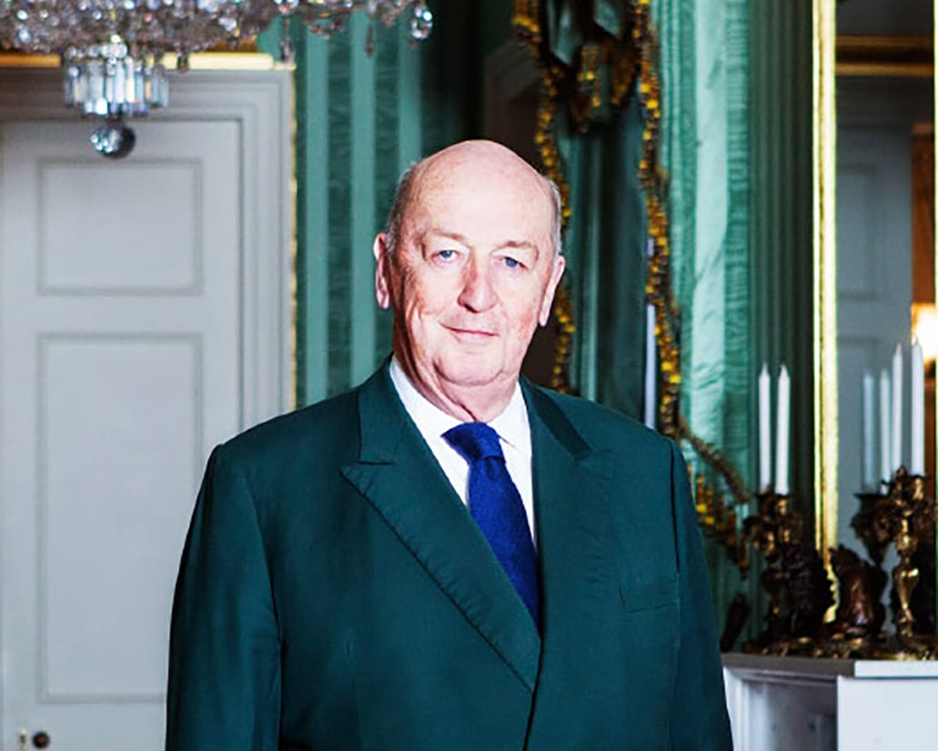 Elderly man in a suit standing in an elegant room with green walls and a chandelier.
