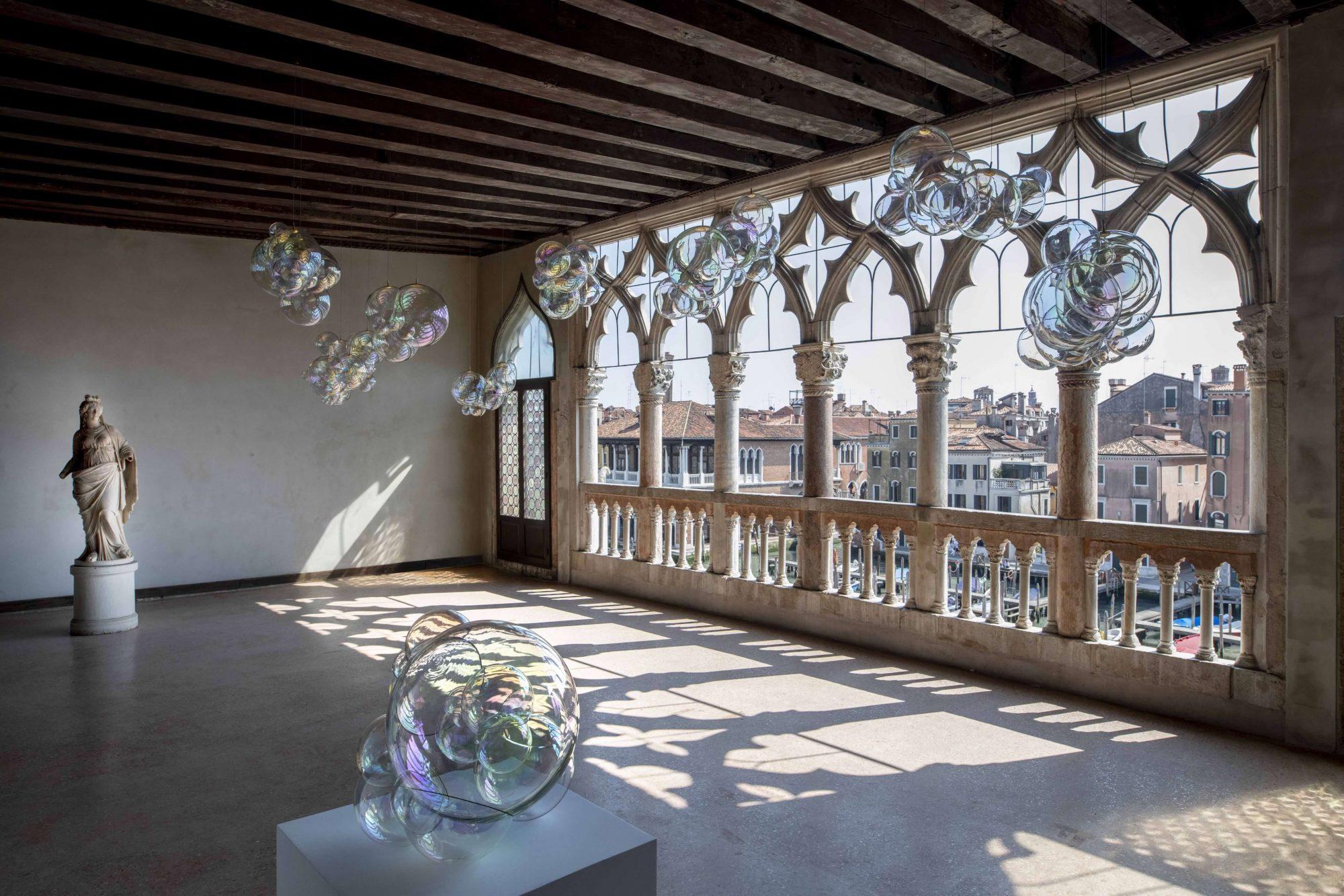 Room with arched windows overlooking buildings, featuring floating bubbles and a statue in a historic interior setting.