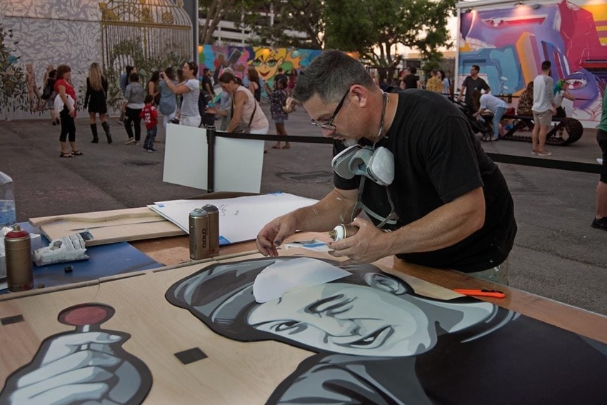 Artist working on a painting at an outdoor art event with colorful murals and people in the background.