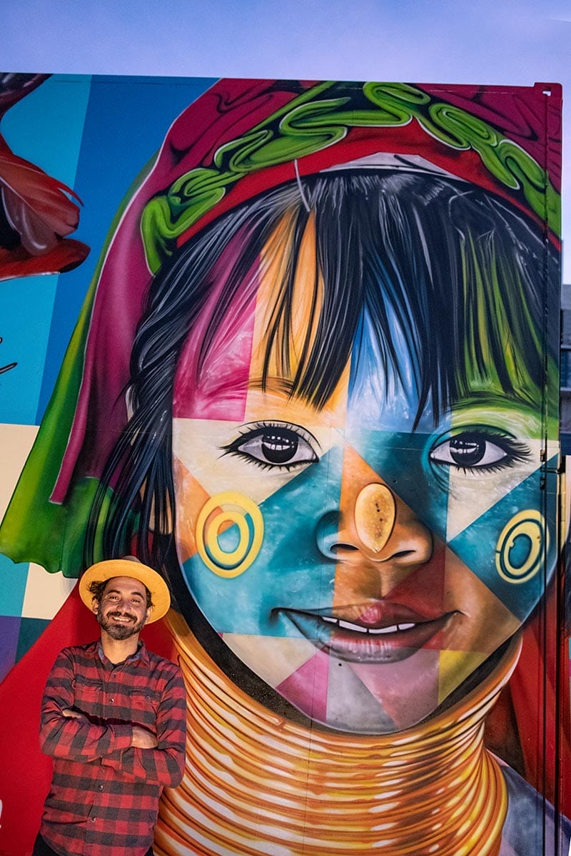 Man in hat stands smiling in front of colorful mural featuring a vibrant portrait of a child with artistic face paint.