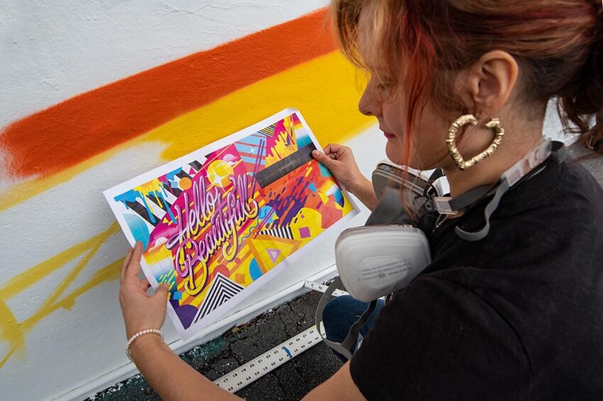 Artist holding a colorful "Hello Beautiful" poster against a partly painted wall with orange and yellow stripes.