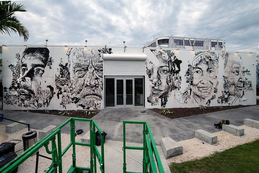 Mural featuring large, detailed portraits on a building exterior under a cloudy sky, with a green lift in the foreground.