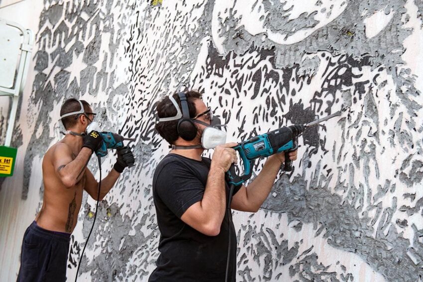Two people using drills to create a detailed mural on a large outdoor wall, wearing protective masks and goggles.