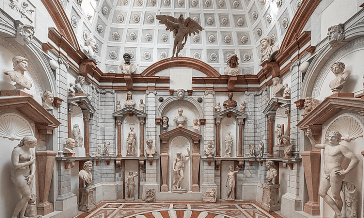 Room filled with classical marble statues, decorative arches, and a detailed ceiling featuring a winged figure.
