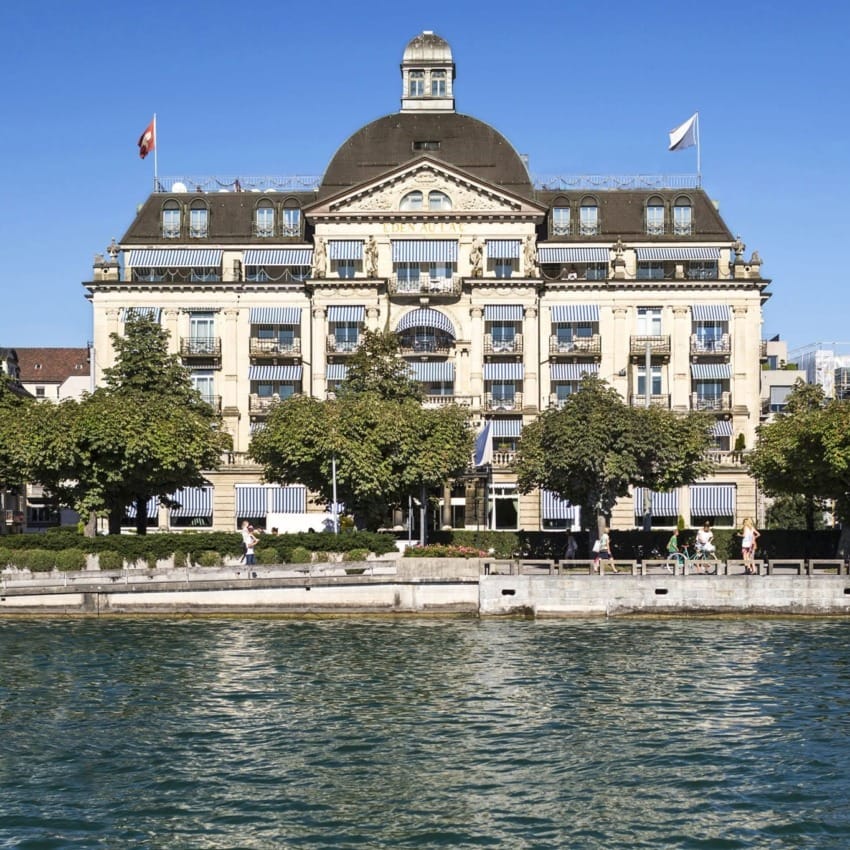 Historic building with ornate architecture by a waterfront, surrounded by trees and pedestrians strolling along the promenade.