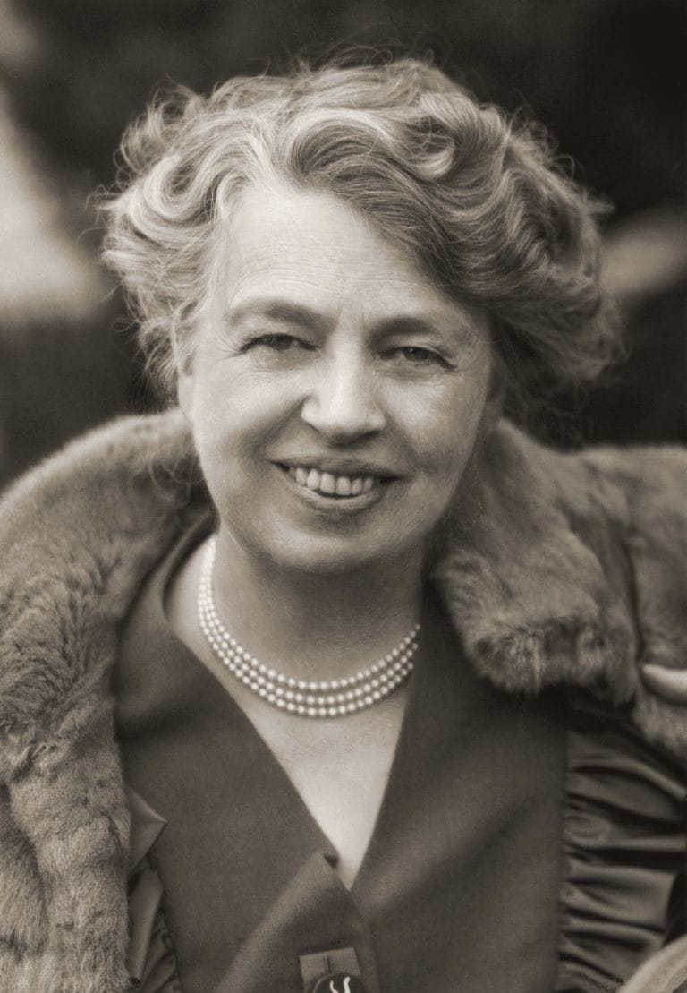 Smiling woman in vintage attire with a fur collar and pearls, sepia-toned photograph, outdoors.