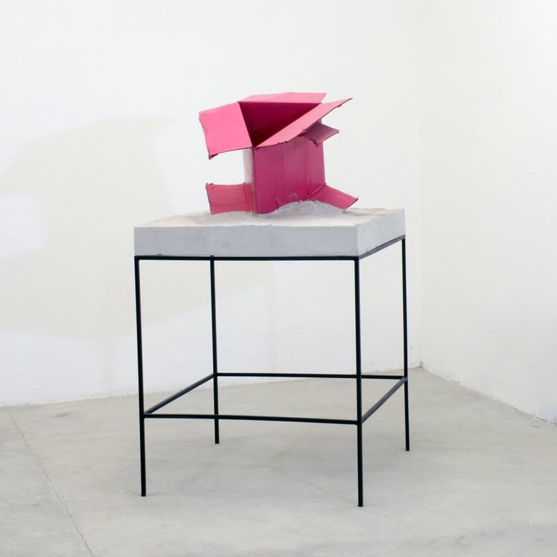Pink geometric sculpture on a gray pedestal with a black metal frame in a minimalist white room.
