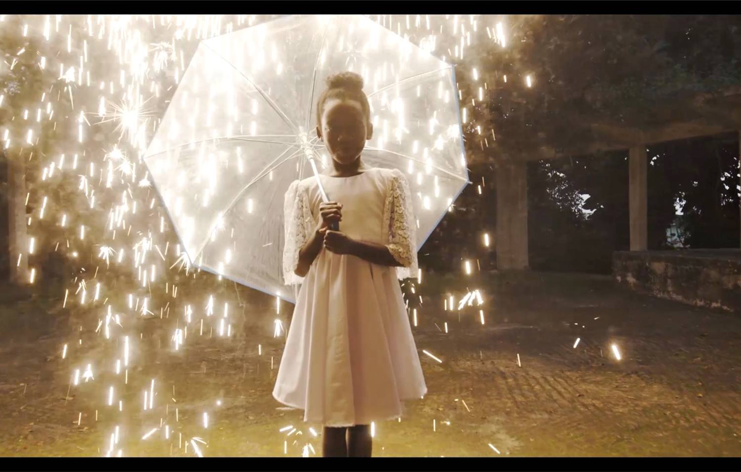 Child holding a transparent umbrella surrounded by sparkling lights in a dimly lit outdoor setting.