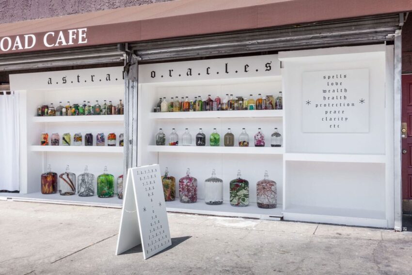 Storefront with shelves displaying various glass jars filled with colorful contents, and a sign listing spells and other items.