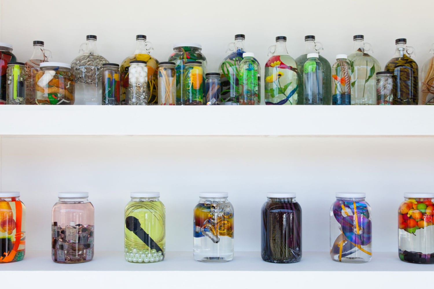Jars of vegetables, fruits, and objects preserved in liquid, displayed on white shelves.