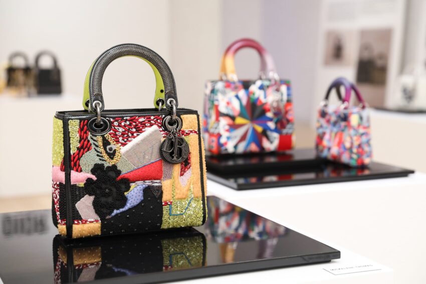 Colorful designer handbags displayed in an art gallery setting with focus on a front bag featuring a textured patchwork design.
