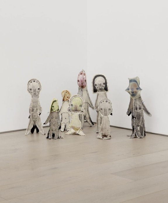 Colorful, abstract sculptures resembling humanoid figures displayed in a corner of a gallery with a wooden floor.