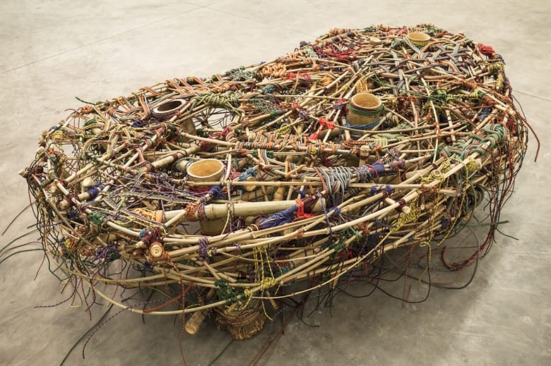 Intricate sculpture made from intertwined sticks, cords, and small objects, resembling an abstract woven seat or structure.