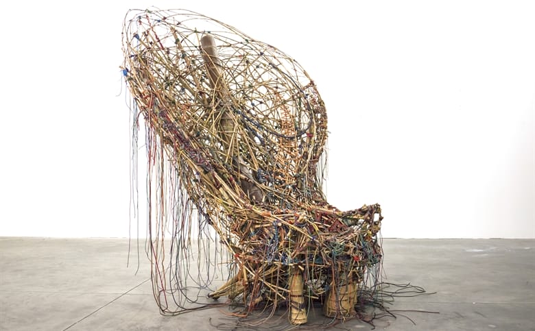Sculpture resembling a chair made from interwoven branches and wires on a concrete floor against a white background.