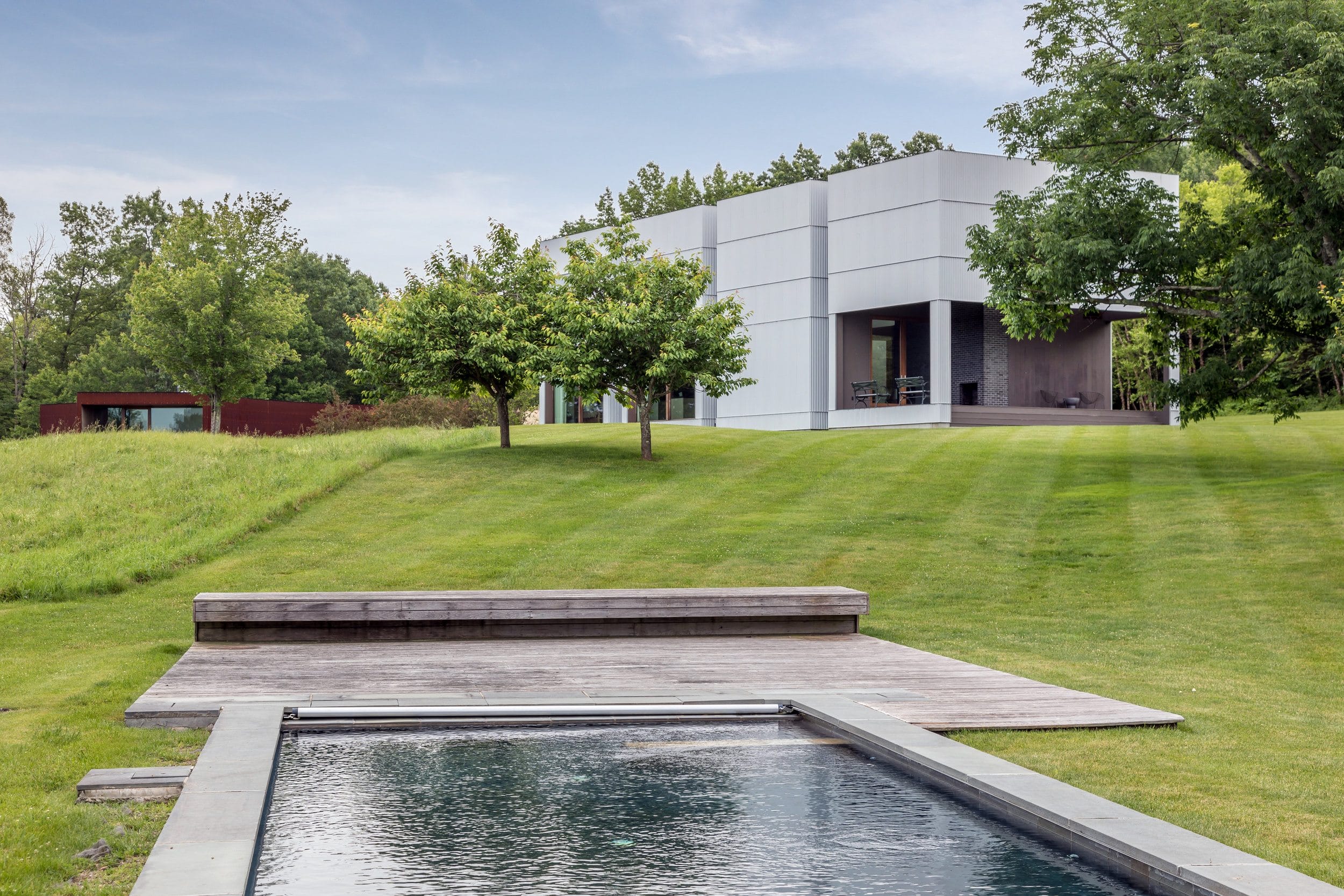 Modern house with geometric design and large windows, surrounded by green grass, trees, and a pool in the foreground.