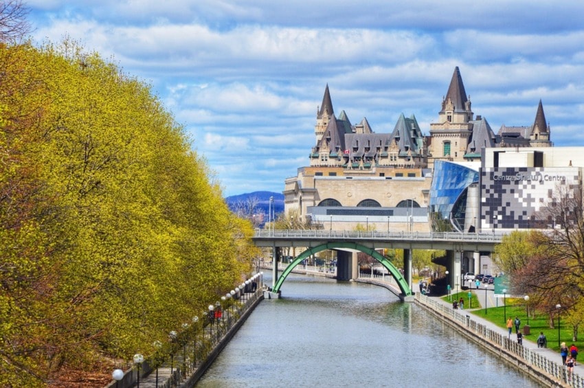 Scenic view of Rideau Canal with green bridge and historic building under a cloudy sky in Ottawa during spring.