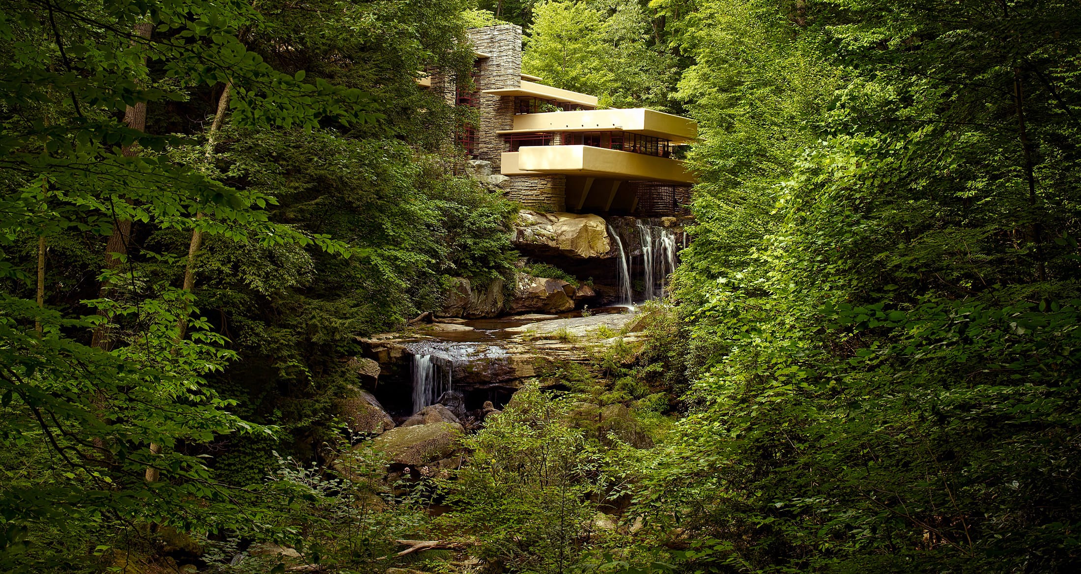 Modern house built over a waterfall surrounded by lush green forest, featuring cascading waterfalls and stone architecture.