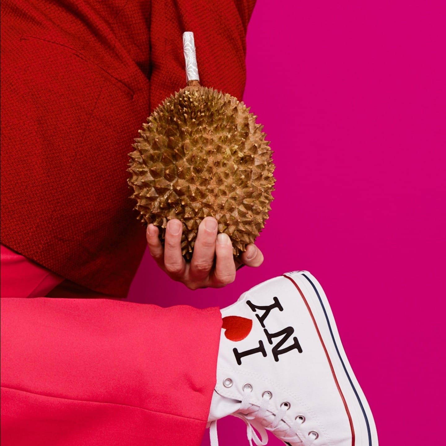 Person holding a durian while wearing red pants and white sneakers with "I ♥ NY" against a pink backdrop.