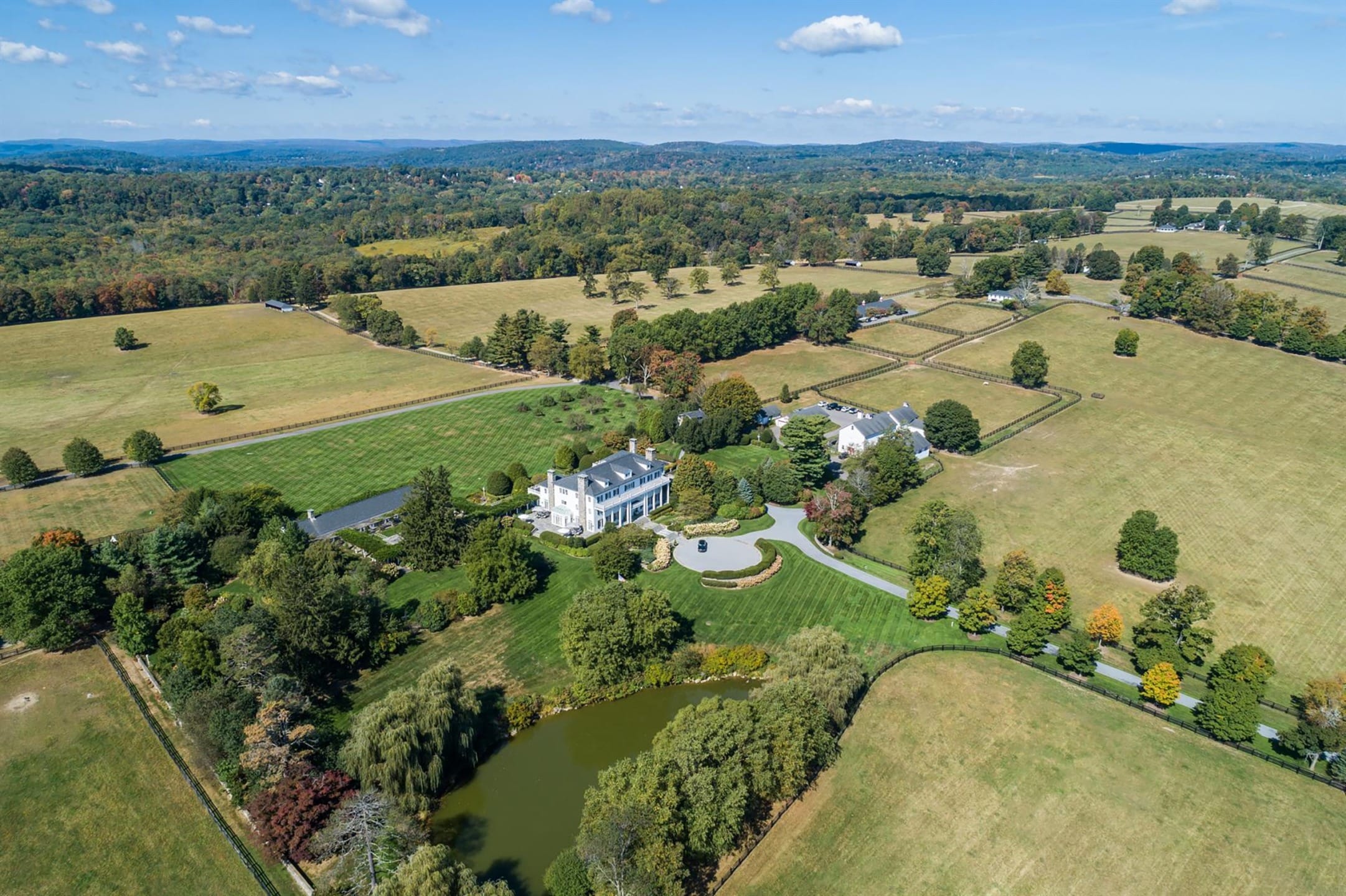 Aerial view of a large countryside estate with a main house, surrounding fields, trees, and a pond under a blue sky.