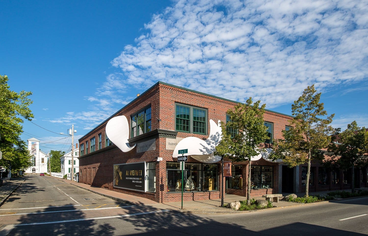 Historic brick building on a sunny day with blue sky, located on a corner street lined with trees in a town setting.