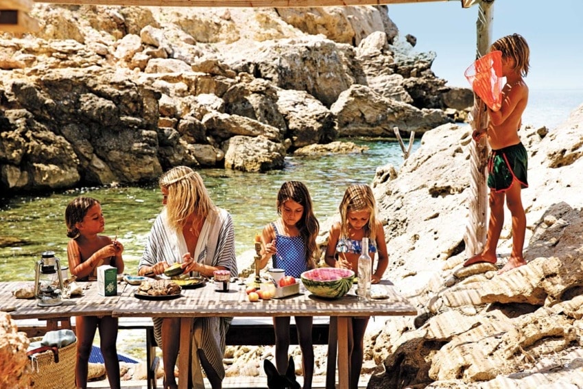 Family eating together at a picnic table near rocky shore, with a child holding a net standing on rocks nearby.