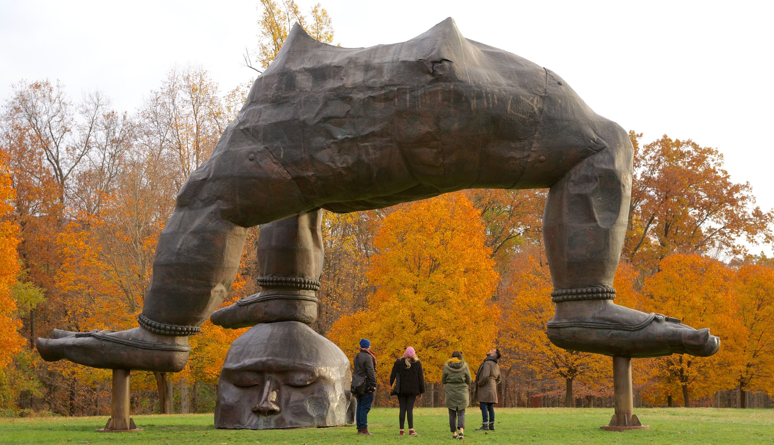 Giant statue resembling a head balancing a pair of legs with four people looking up, surrounded by colorful autumn trees.