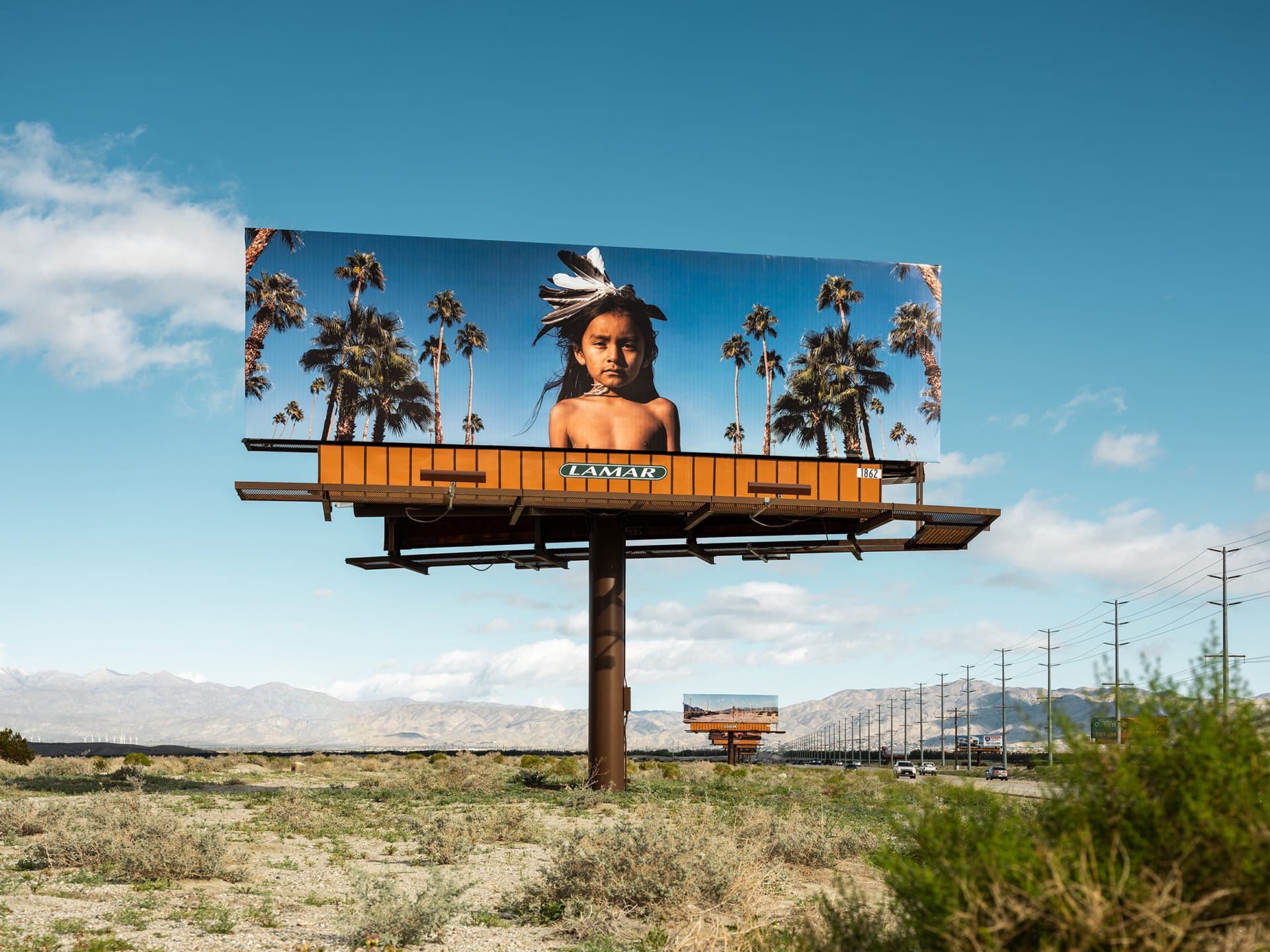 Billboard above desert road featuring a child with palm trees and clear blue sky in the background.