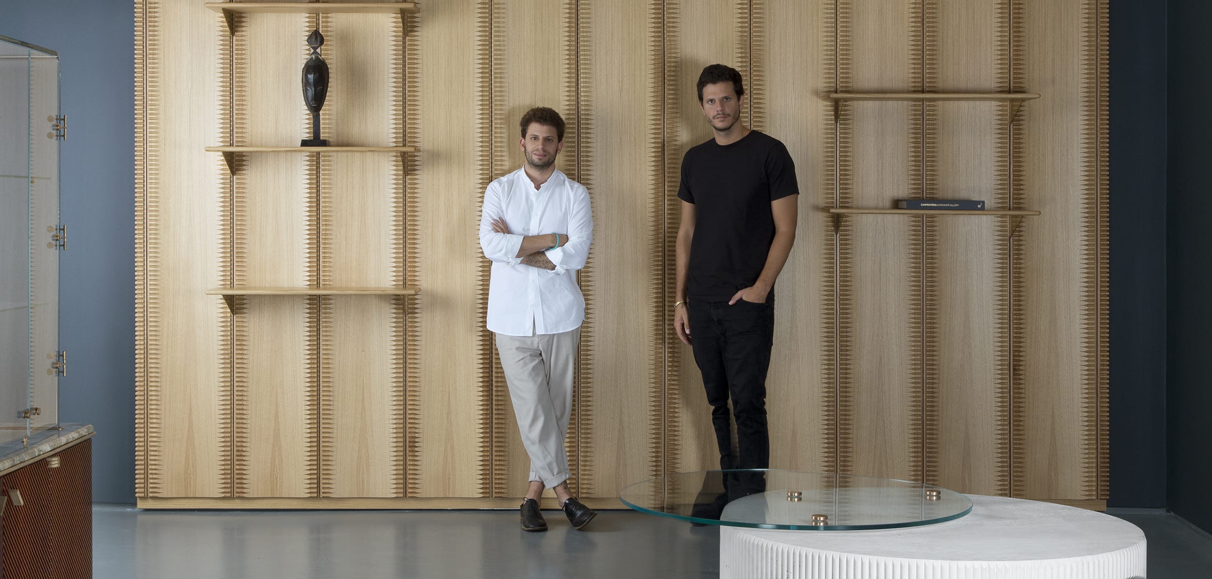 Two men standing in front of a wooden textured wall with decorative shelves and a glass coffee table in the foreground.