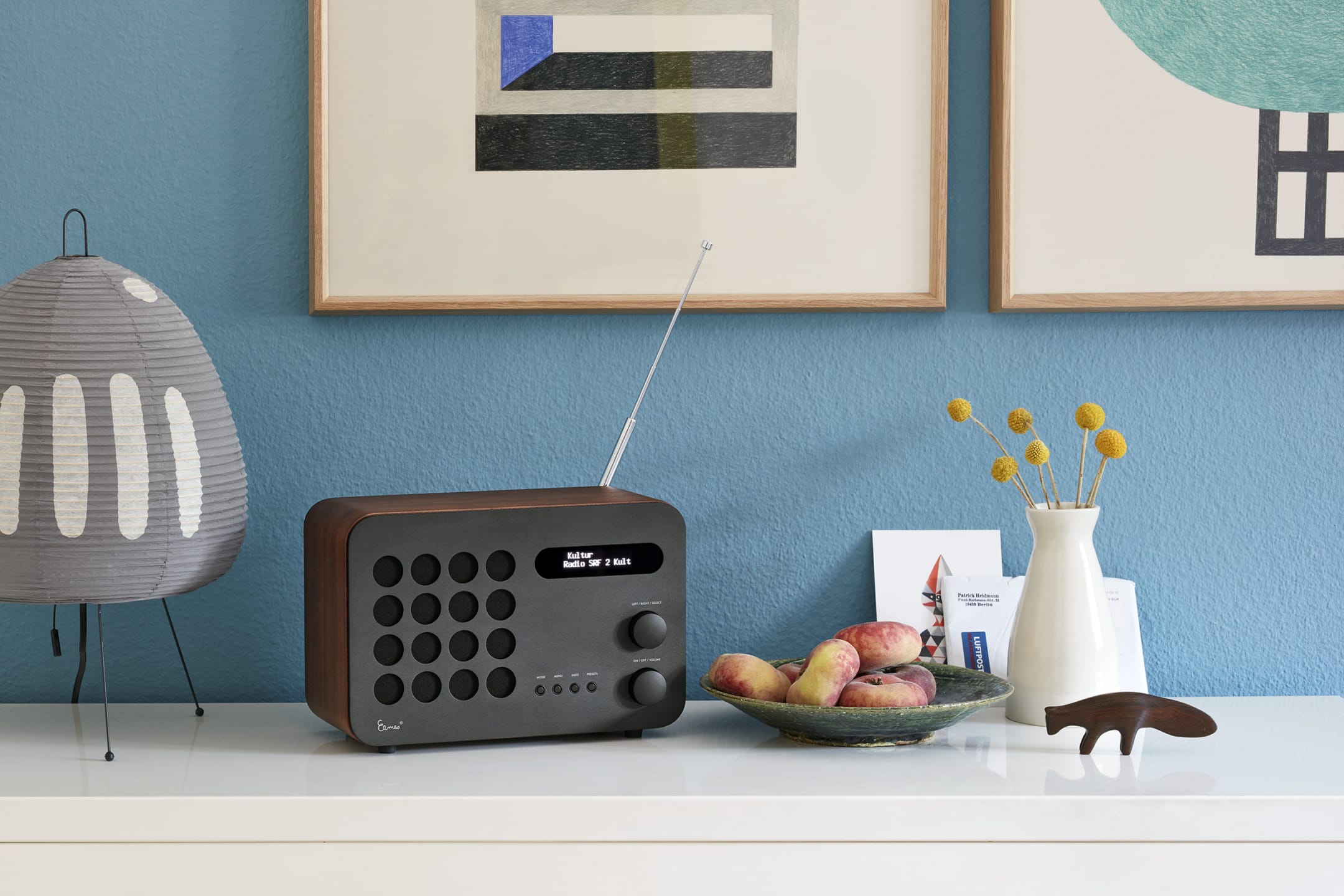 Modern radio on a white shelf next to a bowl of fruit, decorative lamp, and vase with yellow flowers against a blue wall.