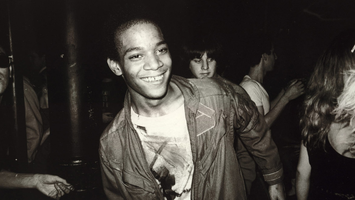 Person smiling at a party wearing a jacket, surrounded by other people in a dimly lit environment.