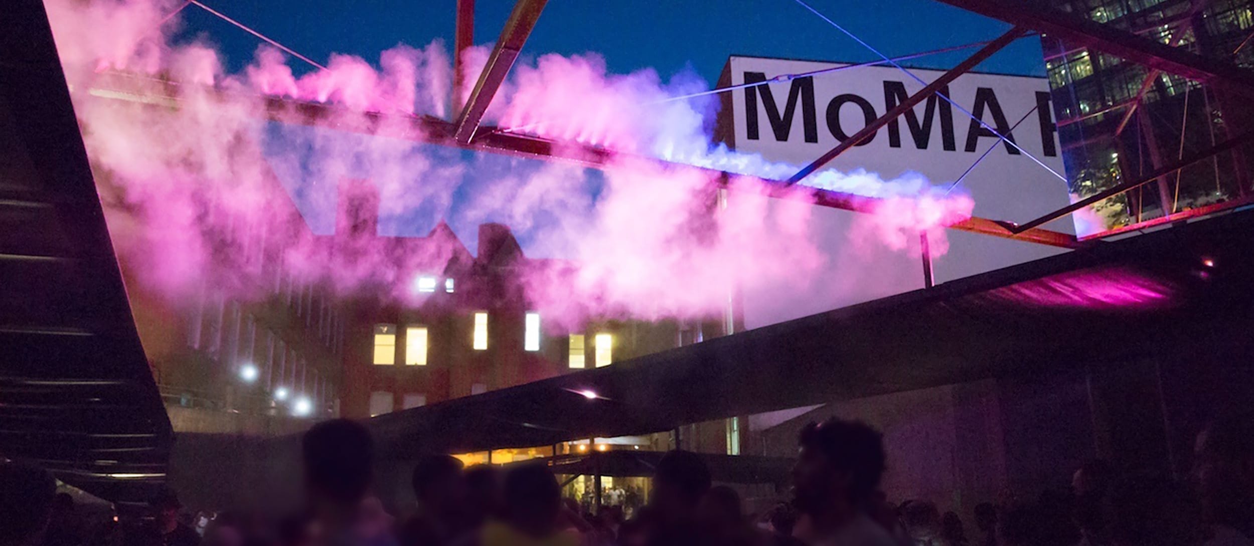 Colorful evening mist installation at an outdoor event near a MoMA museum sign with a crowd gathered below.