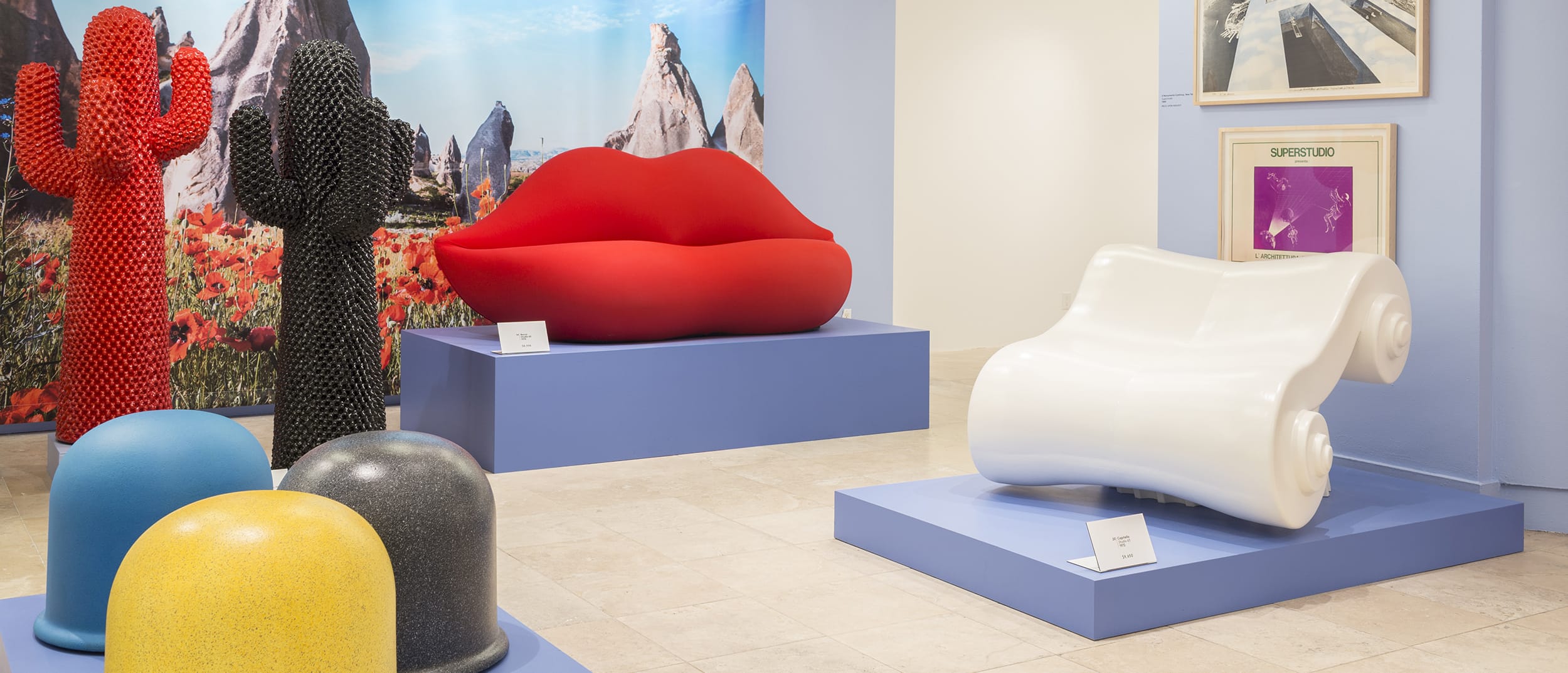 Colorful modern art exhibit featuring sculptures of cacti, large lips, and abstract forms on display pedestals in a gallery.