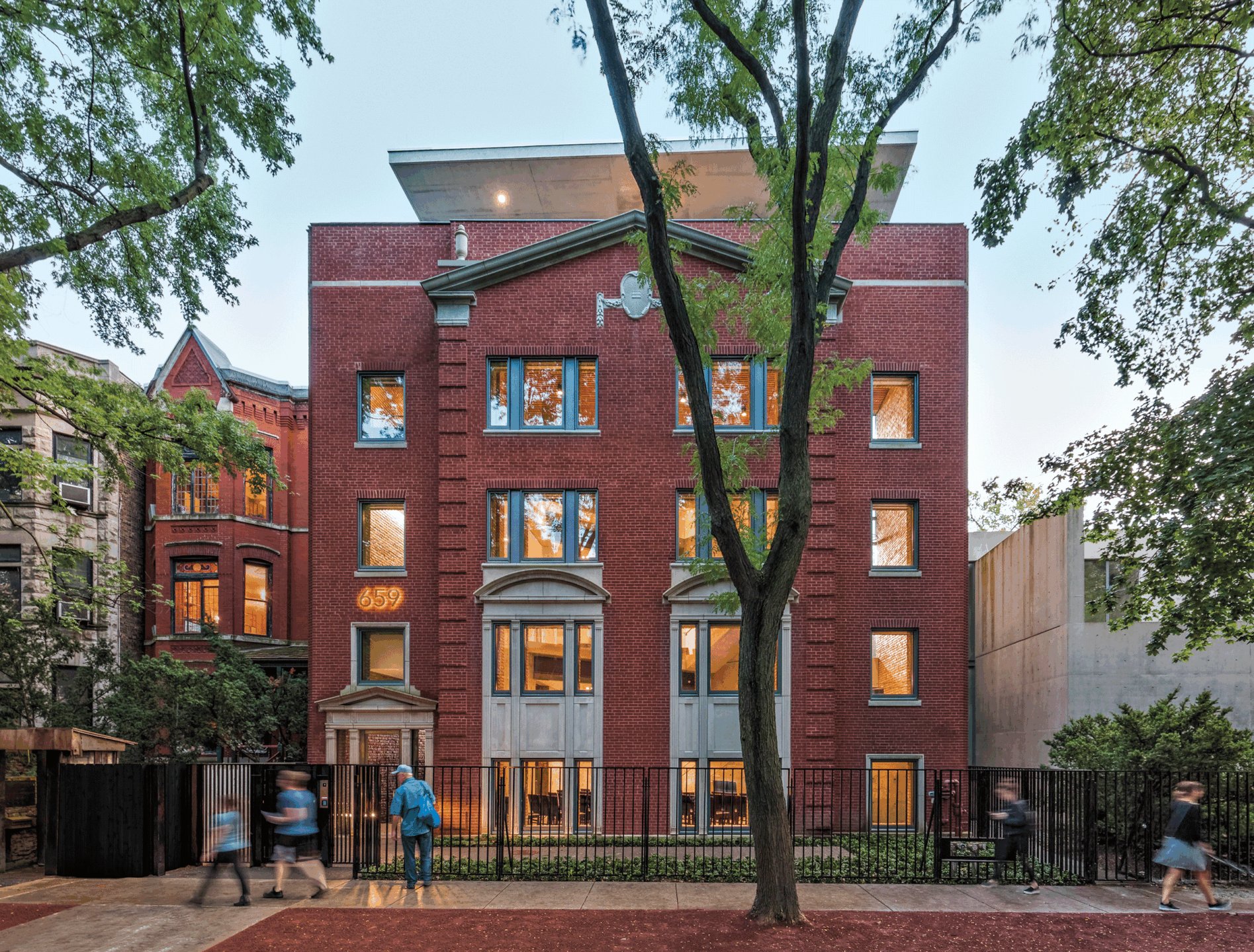 Red brick apartment building with large windows, trees framing the view, people walking by, evening sky in the background.