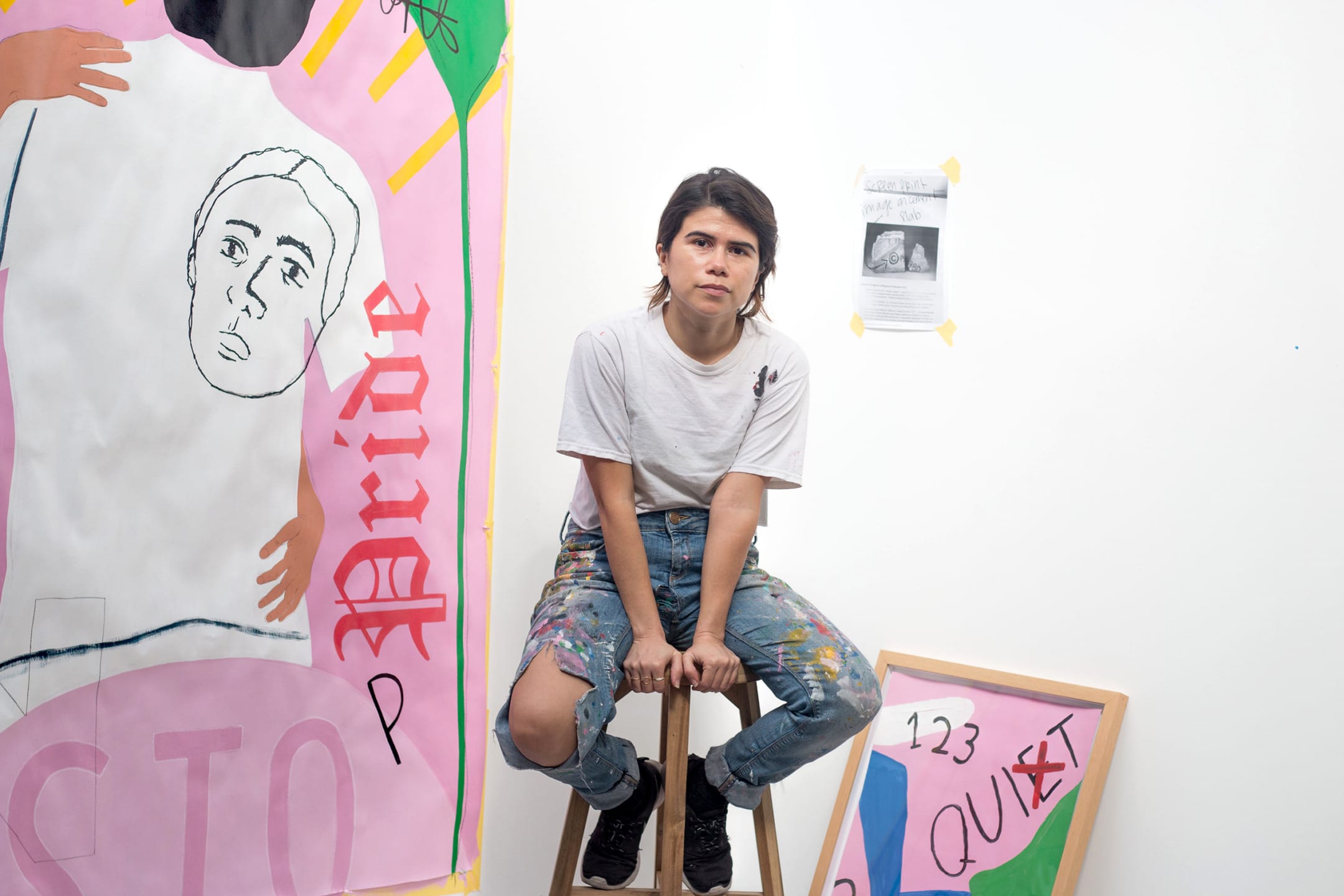 Artist sitting on a stool in a studio with colorful paintings around.