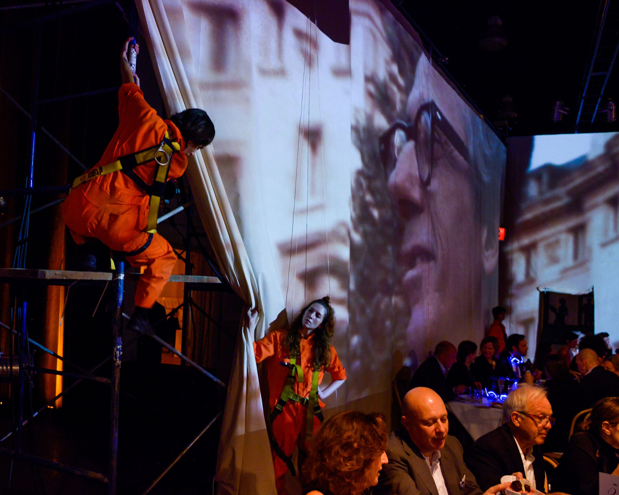 Performers in orange coveralls interact with draped fabric projection backdrop at a lively indoor event with seated guests.