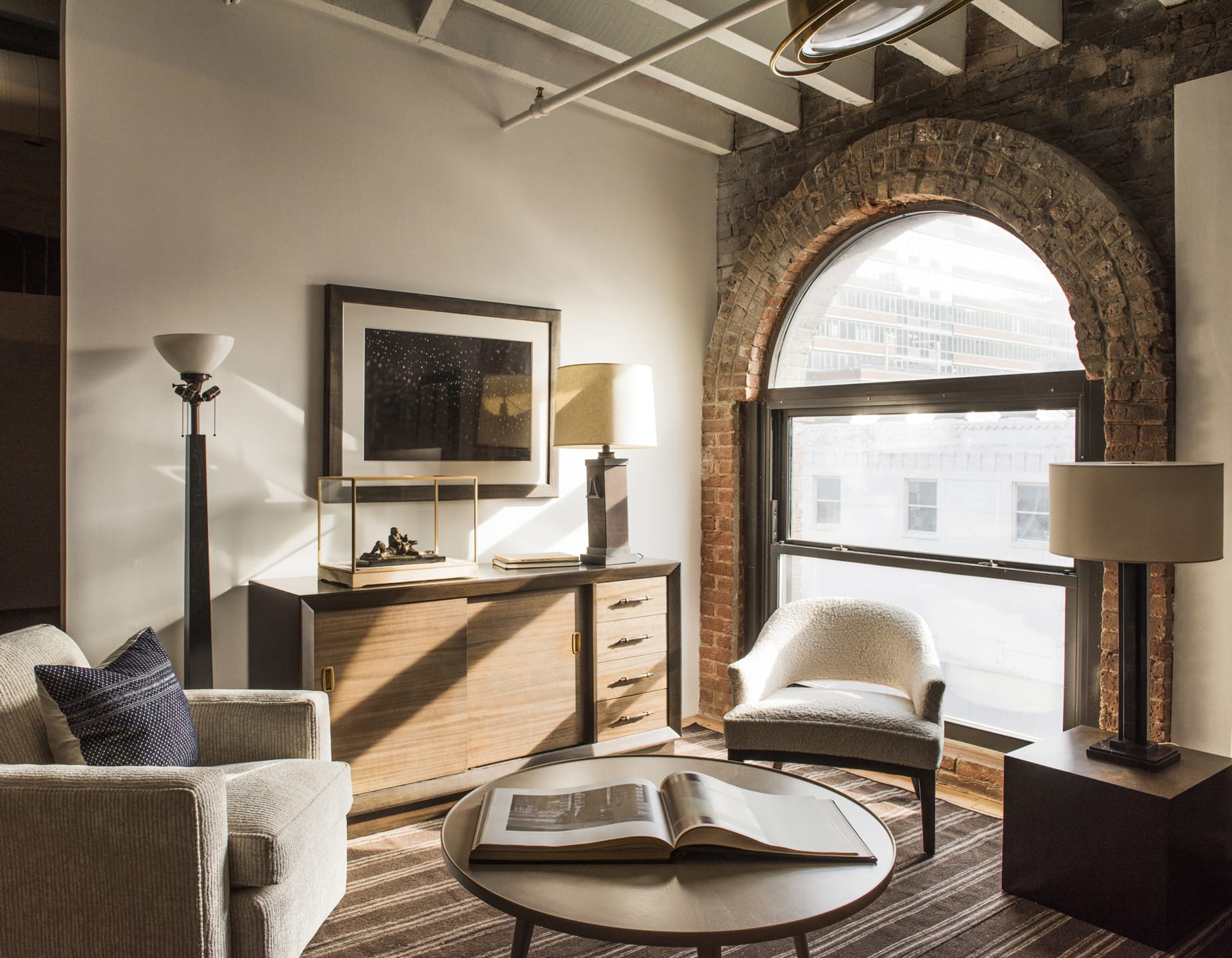 Cozy living room with modern furniture, exposed brick arch window, lamp, and open book on round table.
