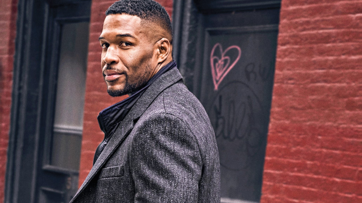 Man in a gray coat stands outside, looking back, with brick wall and graffiti in the background.