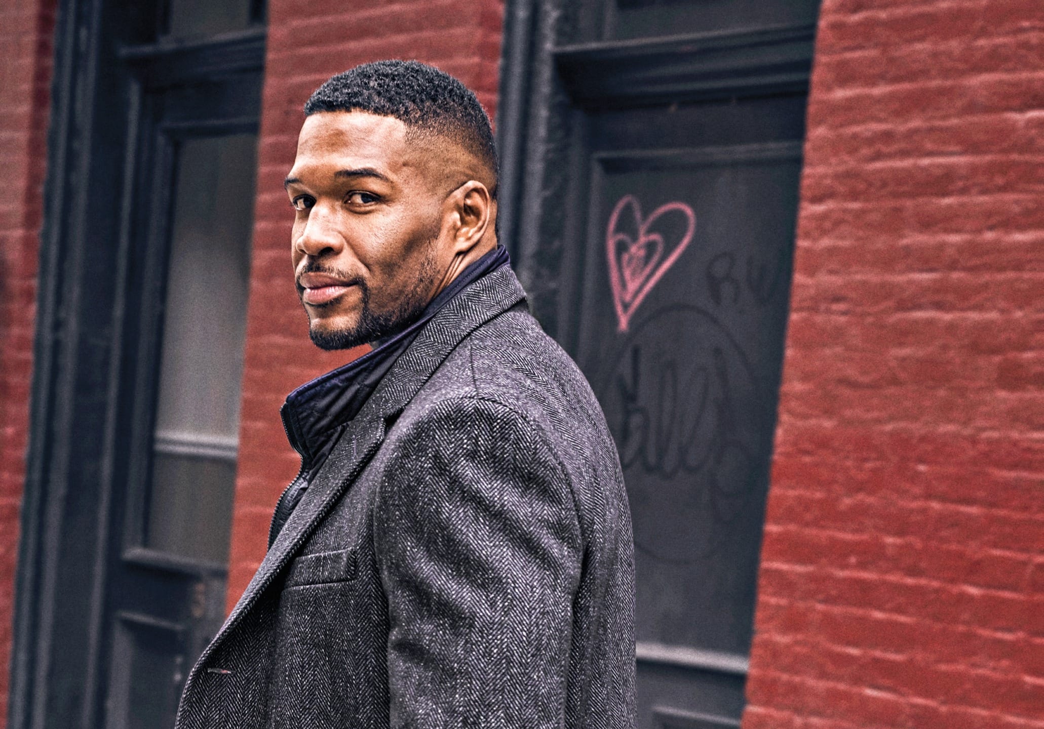 Man in a gray coat stands outside, looking back, with brick wall and graffiti in the background.
