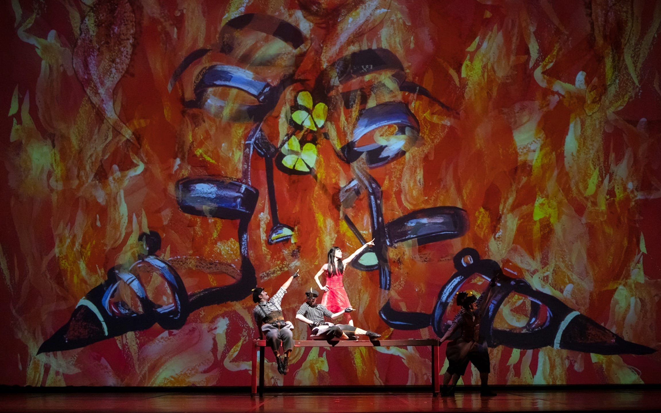 Performers on stage with a vibrant, fiery background and abstract imagery, creating a dynamic and dramatic scene.