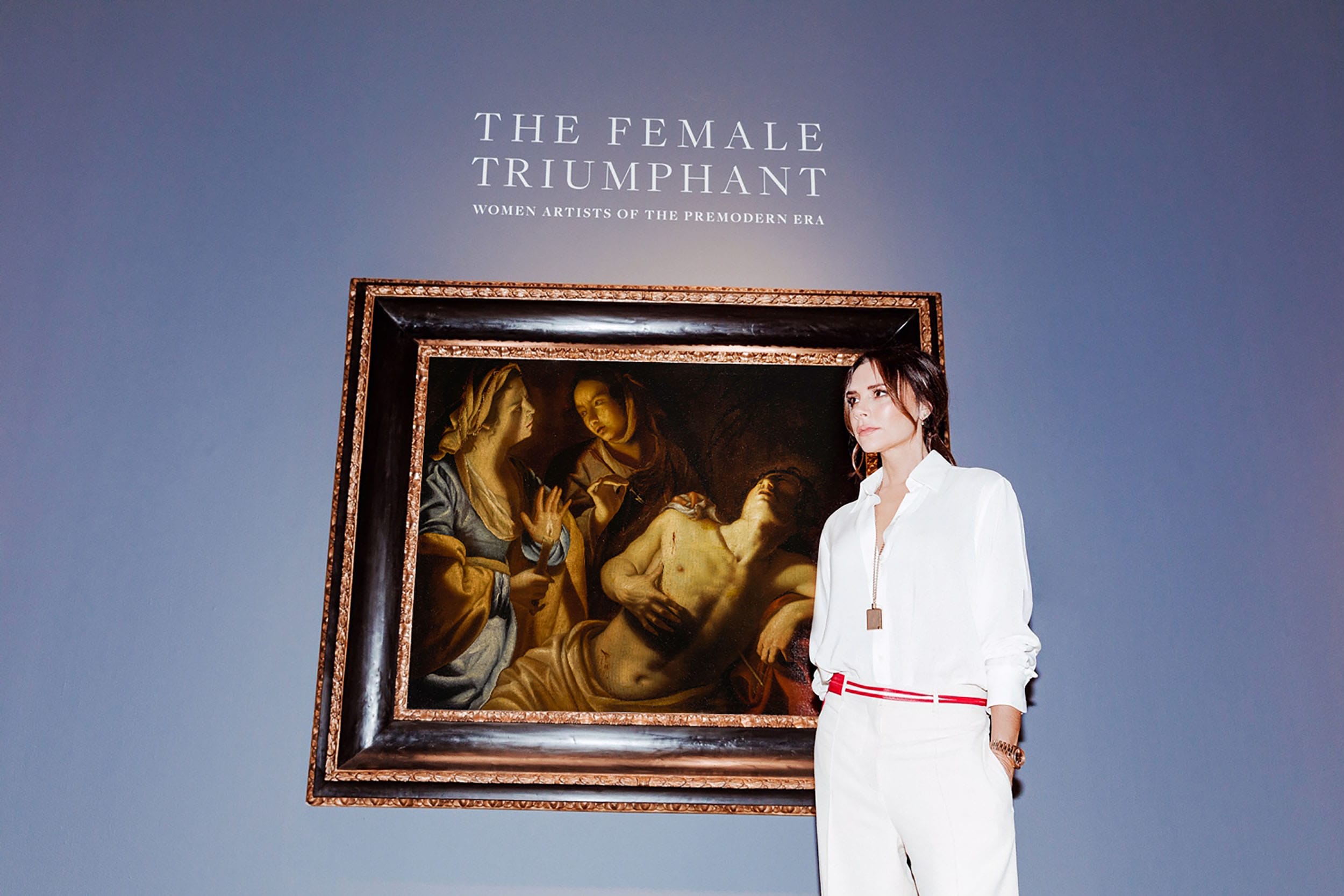 Person in white outfit standing near framed artwork titled "The Female Triumphant" showcased on a blue-gray wall.