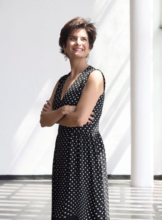 Person in a black polka dot dress standing with arms crossed, smiling in a sunlit room with white walls.