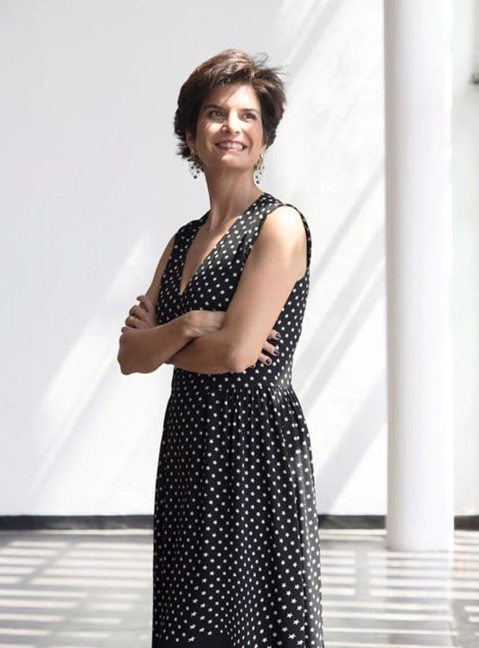 Person in a black polka dot dress standing with arms crossed, smiling in a sunlit room with white walls.